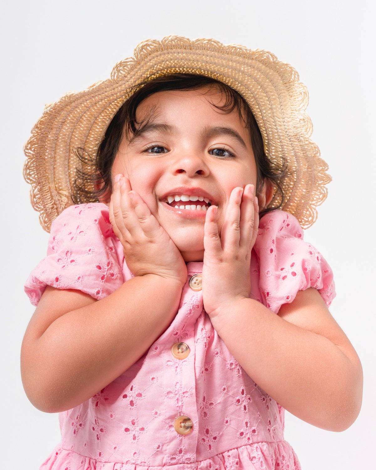 Joyful little girl in pink dress and straw hat, hands on cheeks — children studio portraits Austin & Pflugerville